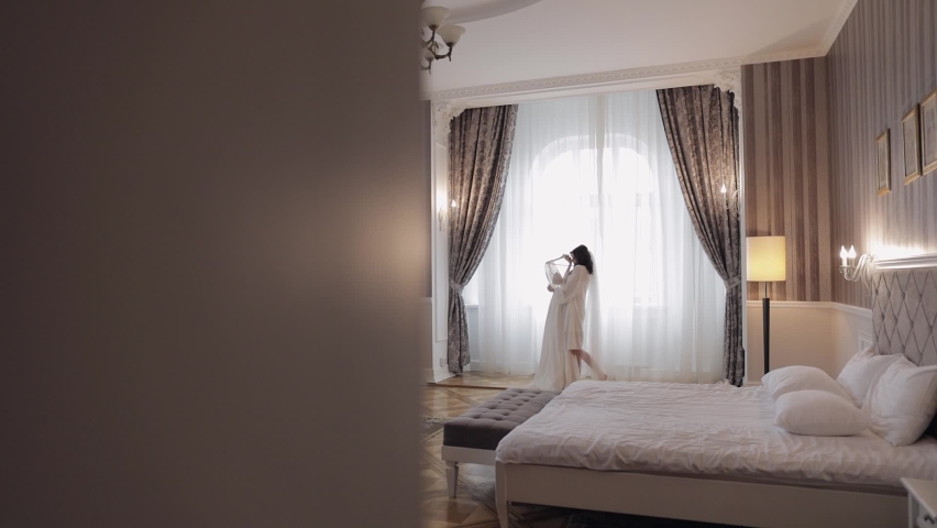 Bride in lingerie standing with her wedding dress. White boudoir dress. Morning preparations indoors. Luxury bride with hairstyle and makeup in a hotel or at home. Woman in white night gown and veil