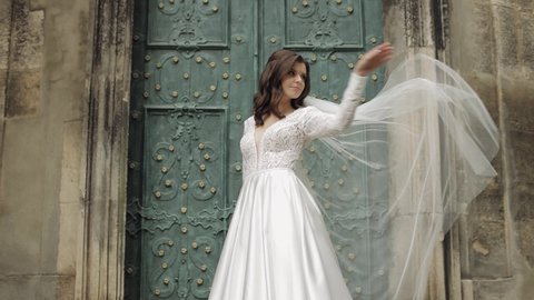 Beautiful lovely stylish bride in white wedding dress and veil near old church waiting for groom. Pretty and well-groomed fashionable woman with fashion hairstyle and makeup. Wedding day. Slow motion