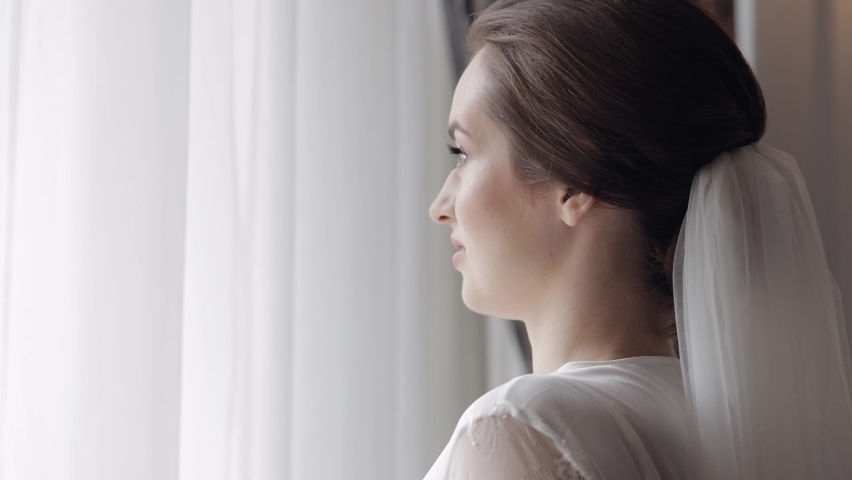 Close-up bride face in white boudoir dress stay near window. Wedding morning preparations. Beautiful lovely woman in night gown, veil. Luxury bride with fashion hairstyle, makeup in hotel or apartment