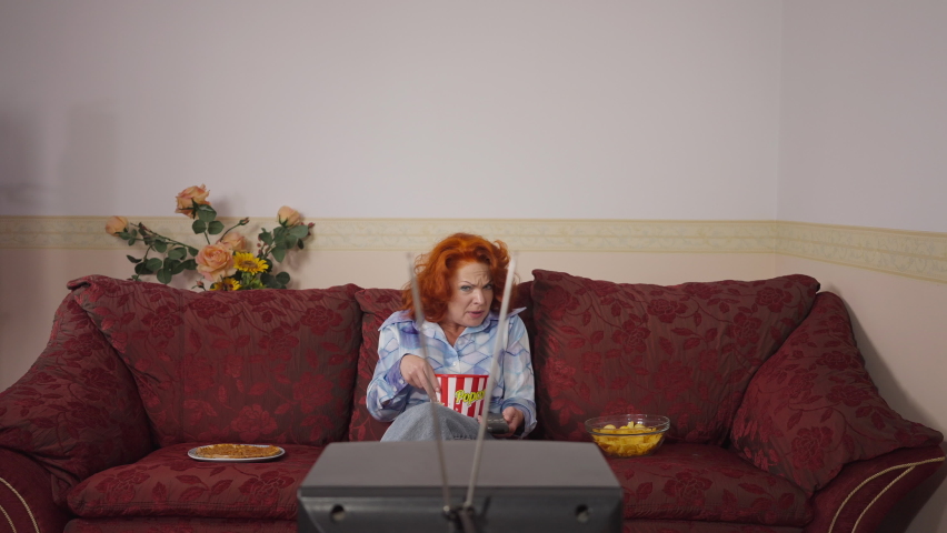 Irritated retro woman hitting vintage TV with antenna sitting back on couch watching movie eating junk food. Portrait of Caucasian lady resting on weekend at home in 1980s 1970s. Slow motion
