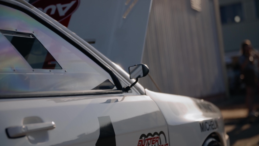Exhibition of white tuning cars parked in an urban setting with an open hood revealing the engine in front of a bunch of people on a sunny day