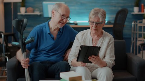 Elder couple looking at tablet screen in living room, enjoying free time together. Retired man with crutches and woman with digital device browsing internet to have leisure activity. Modern people