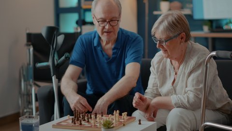 Elder couple with disability playing chess game on board at home. Grandparents having fun with strategy play, moving kind, turn and pawn to do checkmate. Senior people enjoying competition.