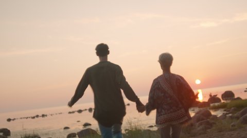 Handheld beautiful footage of authentic millennial couple hold hands and run towards sunset on secluded empty beach. Travellers or lovers hold each other in embrace. Summertime freedom lifestyle