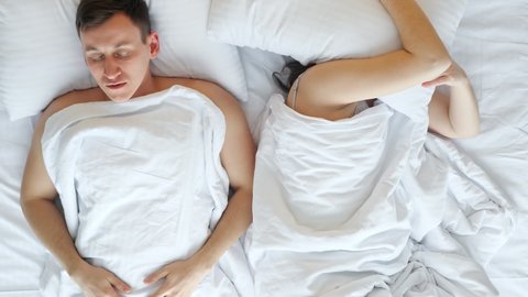 Pregnant brunette woman tries to sleep and wakes up man snoring while sleeping in bed covered with white sheets in bedroom at home, upper view.
