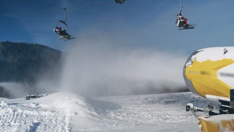 Snow cannon makes artificial snow on background of working ski lift with people, going to mountain slope in sunny day. Snow making system blows water in ski resort, backdrop blue sky. 4k footage