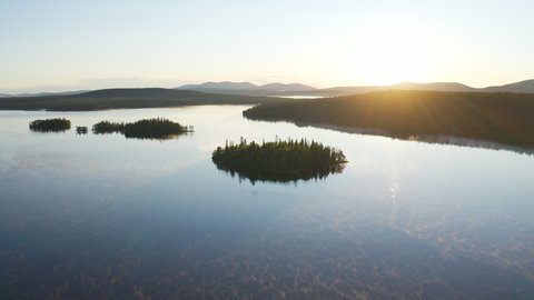 Aerial view high above a blue foggy lake during sunrise showing small islands and mountains in the background as sun is giving golden hour light to the landscape filmed in Lapland Finland.