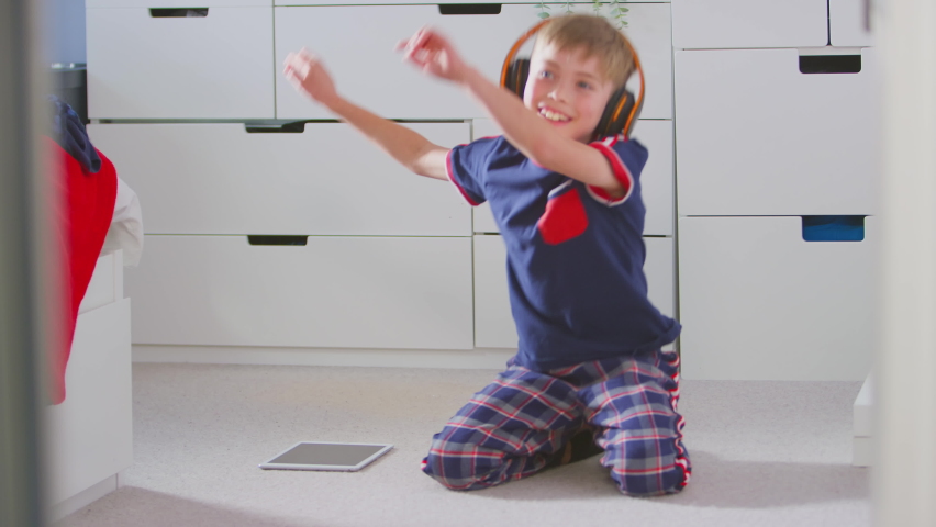 Boy at home in bedroom wearing wireless headphones sitting on floor listening to music and dancing on digital tablet - shot in slow motion