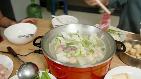 4k Slow Motion, a pot of sukiyaki is on the table with vegetables and pork in the pot. Hands are scooping pork into the pot waiting to be cooked to eat.