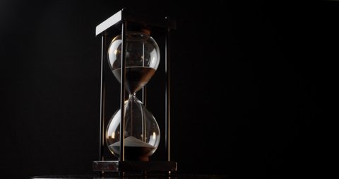 Rotation of sand timer clock with sand flowing from through hourglass on black background.
