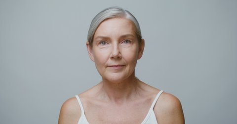 Professional elderly model of European appearance without makeup, with gray hair gathered in a low ponytail, posing looking at the camera, touching her face with her hands. 4K video, red komodo
