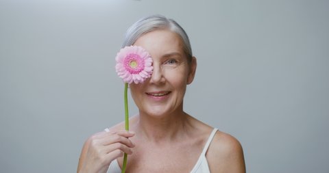 A senior Caucasian model with no makeup, gray hair pulled back, posing with a gerbera flower, stroking her face and looking at the camera with a wide smile. 4K video, red komodo