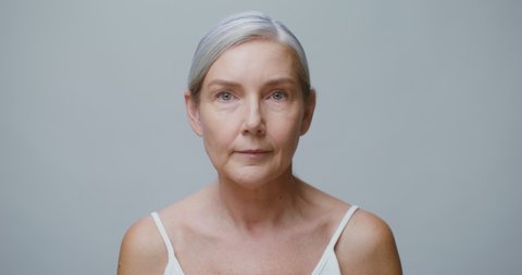 An older fair-skinned model with pulled gray hair applies moisturizer to her face while smiling at the camera, standing against a solid-colored studio background. 4K video, red komodo