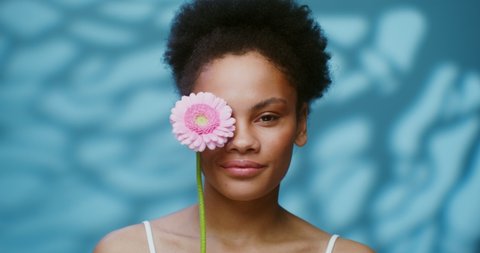 A young African American model with short curly hair poses looking at the camera, running a pink gerbera flower along her face and smiling broadly. 4K video, red komodo