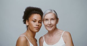 An elderly gray-haired model and a young African American woman pose together smiling looking at the camera while standing in front of a monochrome studio background. 4K video, red komodo