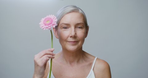 An aged caucasian model, without makeup, with gathered gray hair, posing with a gerbera flower, stroking her face and looking at camera, against a monochrome studio background. 4K video, red komodo