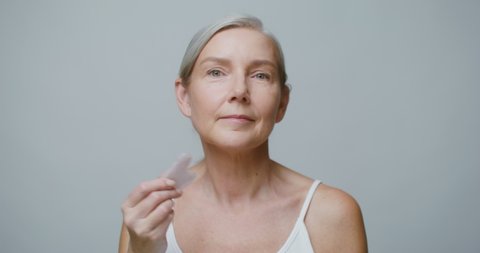 A professional elderly Caucasian model without makeup, with gray hair pulled back in a low ponytail, doing a facial massage using a Gua Sha stone while looking at the camera. 4K video, red komodo