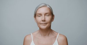An elderly Caucasian model with no makeup, gray hair pulled back, doing a facial massage using a jade roller and looking at the camera, against a monochrome studio background. 4K video, red komodo