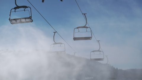 4k empty ski lifts against blue sky in motion, artificial snow sprayed by snow cannon in sunny winter, bottom view. Artificial snow cloud, backdrop of working chair lift without people in ski resort