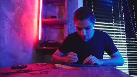 Young craftsman making dream hunter in dark workshop. Dreamcatcher creation. Artisan at work in futuristic art studio illuminated by neon red and blue lighting.