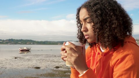 Panning shot of beautiful mixed race African American girl teenager young woman wearing orange hoodie, drinking takeout coffee by a harbour looking sad or thoughtful