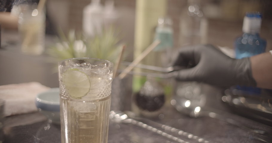 Cocktail series. Bartender preparing a Peruvian cocktail called Pisco Sour, in a bar with a relaxed atmosphere in slow motion, add ice, lime and mix the drink wearing black gloves as biosecurity 
