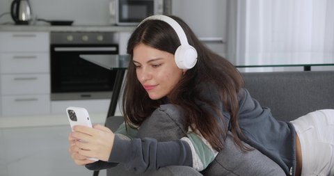 teenage girl listening to music wearing headphones relaxing on sofa texting using smartphone browsing online playing mobile games enjoying sharing messages on social media