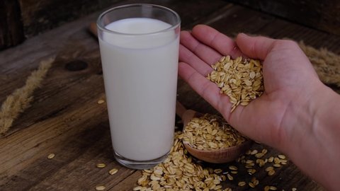 Female hand pouring whole grain oats near glass of vegetable oat milk on wooden background