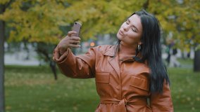 Close-up young hispanic woman stands in autumn park holding phone in hand girl blogger posing photographed on mobile camera make foto for blog using smartphone lady vlogger makes online streaming