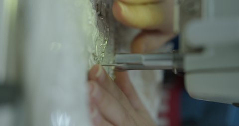 seamstress on a sewing machine embroider shiny fabric with a needle in production. A woman sews a wedding dress on an automatic machine. Vertical video. Close-up