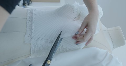 a woman holds big black scissors in her hands and cuts a white fabric on a corset that stands on a manikin against the backdrop of the production of wedding dresses. Cutting the dress on the manikin.