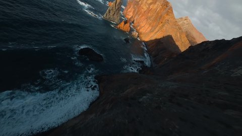 Flying Over Madeira Rocky Coastline alongside Cliffside with Blue North Atlantic Ocean Water Towards the Sun During Sunset. Bird Eye View 4K Drone Footage. Soar Above Sea Coast From Up in the Air