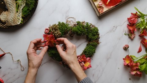 DIY idea, Valentines day decoration. Female hands makes flower arrangement, decorates heart shaped floral wreath for Saint Valentines day. Table top view, POV.