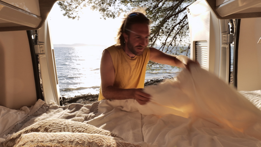 Young man travelling by camper van makes his bed in the morning with beautiful sea view, sunrise 