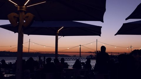 Lisbon, Portugal - January 7, 2022: Silhouette of diners sitting at a cafe overlooking the iconic 25 April Bridge in Lisbon at sunset