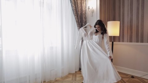 Bride in lingerie dancing with her wedding dress. White boudoir dress. Morning preparations indoors. Luxury bride with hairstyle and makeup in a hotel or apartment. Woman in white night gown and veil