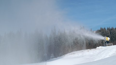 Snow cannon blows artificial snow on mountain slope, background of forest, blue sky in ski resort on sunny day. Snowgun makes snow of water in cold winter. 