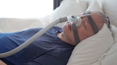 Middle age man with sleep apnea health condition wearing CPAP hear gear connecting to air tube, laying down to sleep in his bed