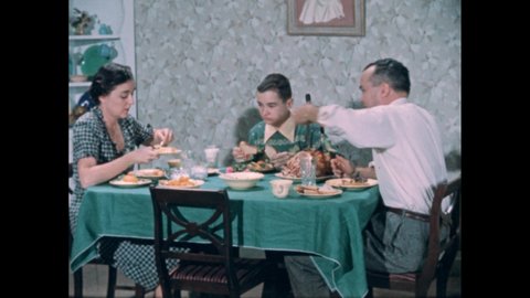 1950s: Family eating dinner superimposed over a table. Man in shirt sleeves and gestures with his fork. Boy grabs bread. Man reaches across table to get butter. Blur. Man wears a coat. Restrained.