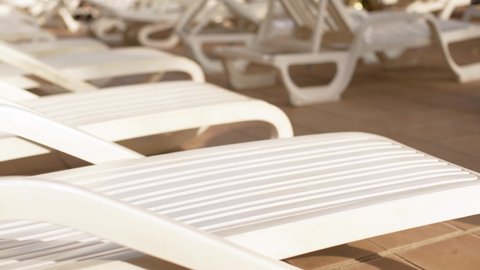 Empty white plastic sunbeds at holiday resort near pool, closeup detail, blurred people moving in background