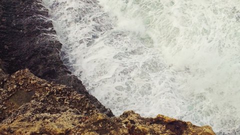 Sea crashing on rocky shore, green water splashing and creating white foam, view from above, slow motion video