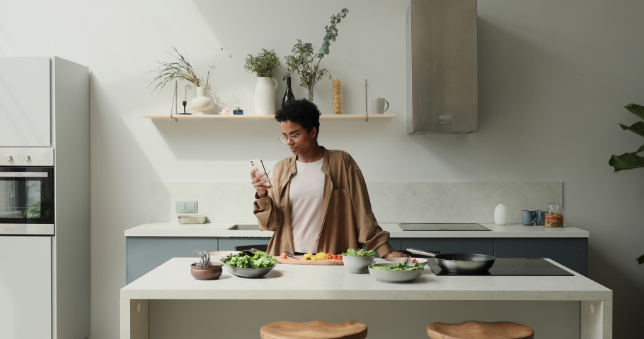 Young African woman stand in kitchen distracted from vegetable salad preparation use smart phone, smile chat online, enjoy modern tech. Lifestyle, on-line recipes, food delivery services user concept Royalty-Free Stock Footage #1085053453