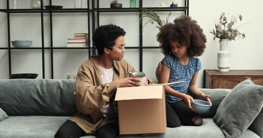 African mum her 10s daughter sit on sofa at home with parcel box, unpack diverse kitchenware, bowls and cups. Satisfied clients family of e-commerce online services, modern dinnerware store ad concept Royalty-Free Stock Footage #1085053459