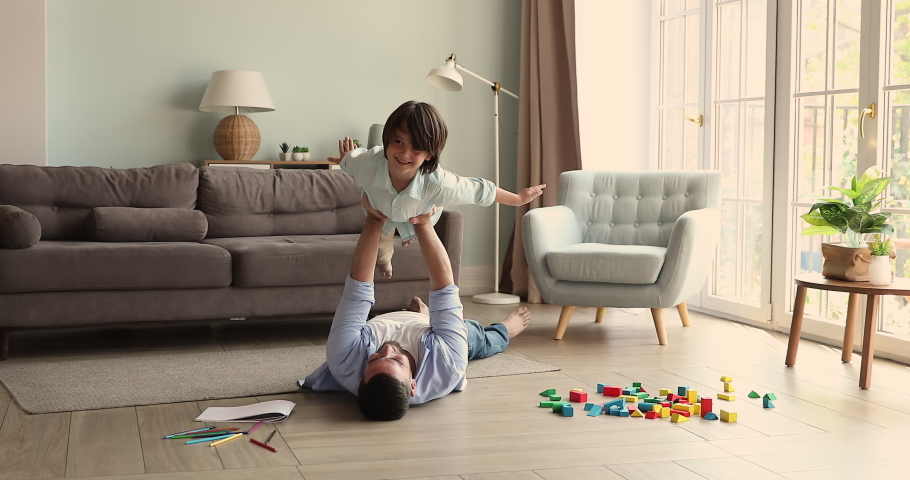 Caring single dad lie on warm heated floor at living room hold cute smiling little son in air on outstretched arms. Happy small child boy pretend airplane superhero flight with help support of father Royalty-Free Stock Footage #1085053504
