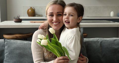 Beautiful woman holding white bouquet fresh tulips hugs little 4s daughter look at camera. Loving family celebrate Happy Mother Day or birthday feeling love and affection, life events congrats concept