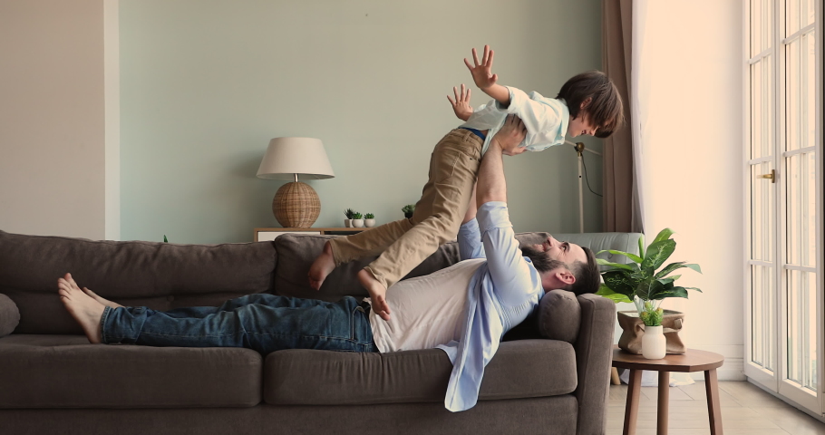 Loving father lie on back on couch play plane with excited son preschooler raise little boy in air keep balance imitate flight. Dad lift child up on strong hands enjoy funny exercise together with kid Royalty-Free Stock Footage #1085053555
