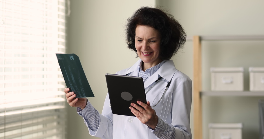 55s woman therapist practitioner in uniform holds xray medical procedure results and digital tablet smile feels satisfied by prescribed treatment of patient. Medicare, diagnostics, modern tech concept Royalty-Free Stock Footage #1085053558