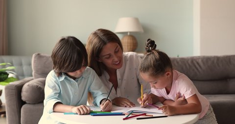 Young woman nanny teacher art therapist engage two little kids elder boy younger girl in creative activity. Focused mother talk play with children help to paint pictures with pencils in bright colors