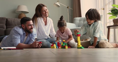Smiling happy parents with small daughter son gather together on warm heated floor play creative game. Foster mom dad two adopted kids construct fantasy buildings from wood blocks at cozy living room