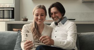 Attractive cheery couple sit on sofa look at cellphone screen laugh watch video, enjoy new cool mobile app, make videocall enjoy online content or virtual meeting. Fun use modern wireless tech concept